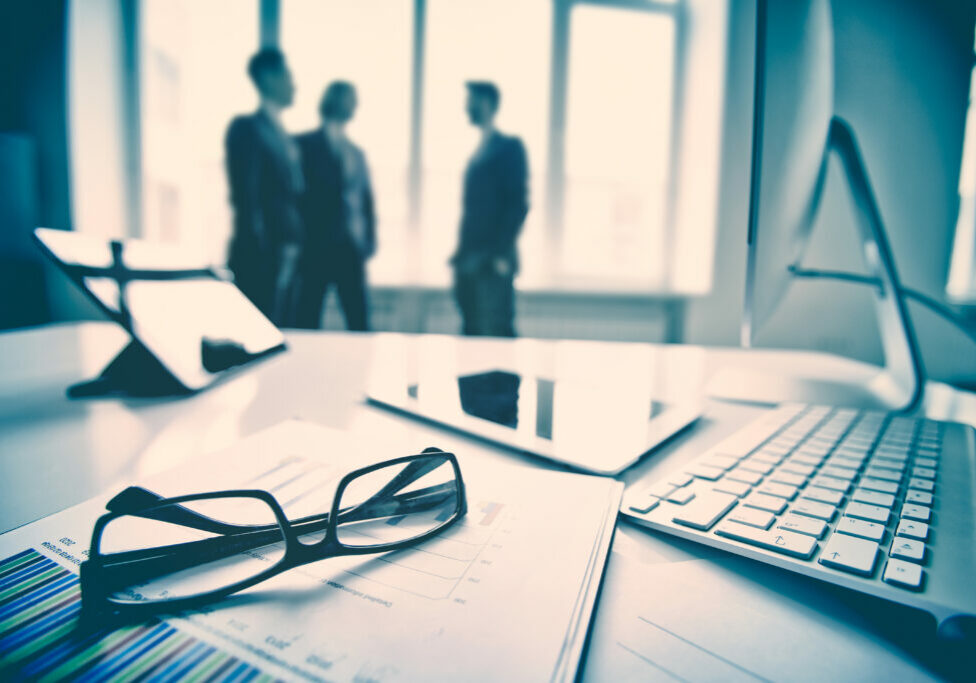 Modern devices, documents, eyeglasses on the foreground, unrecognized businessmen on the background